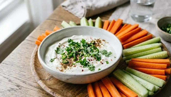 A bowl of thick white curd dip garnished with green chillies and coriander, surrounded by fresh cucumber and carrot sticks on a platter.